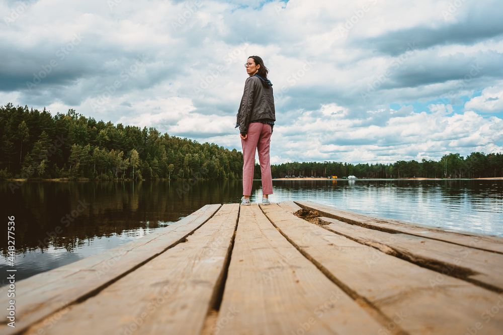 The tourist girl stands with her back on the wooden pier of the forest lake and turns around