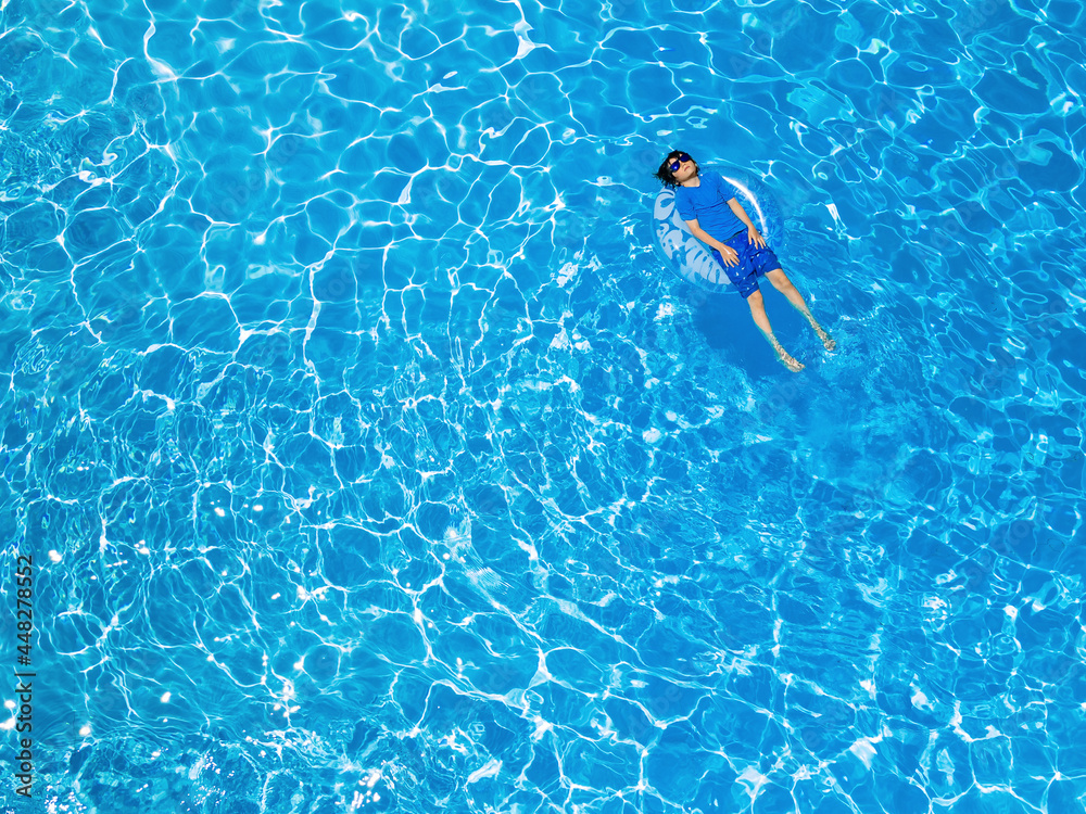 Aerial drone view of  a young boy of ten years old wearing sun protection t-shirt and sunglasses is lying on the inflatable ring in the swimming pool. Summer holidays concept, copy space on the left.