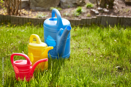 Drei unterschiedliche Gießkannen in blau, rot und gelb stehen im Gras. Die Gartenarbeit ruft für die ganze Familie.