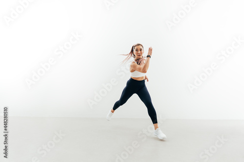 Young sportswoman running forward indoors. Muscular female exercising against a white wall.