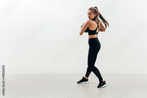 Young muscular woman doing exercises with a medicine ball against a white wall