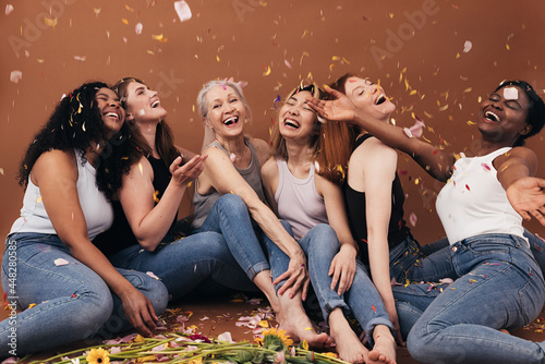 Group of six laughing women of different ages sitting under falling flower petals. Multi-ethnic smiling females having fun in studio while sitting on brown background.