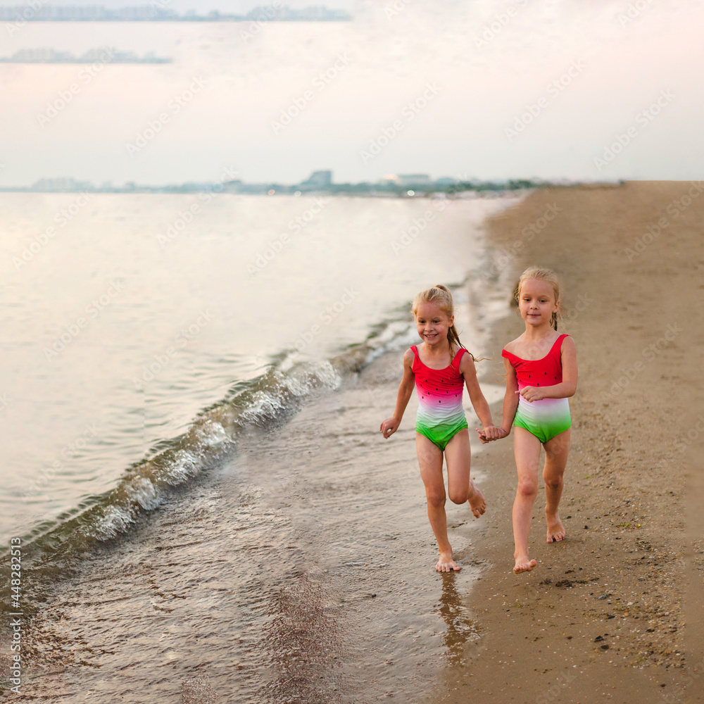 two little girls sisters run in summer in swimsuits with a pattern of watermelon along the seashore