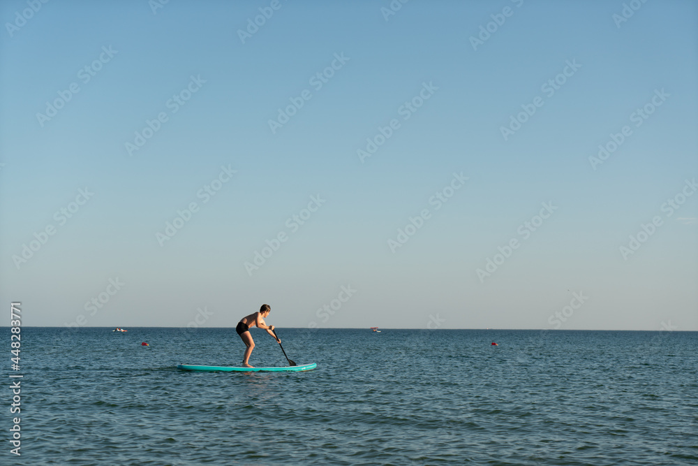 A 12 year old boy learns to stand on a SUP board in the sea near the shore.