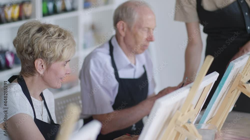 Side view of a happy senior people smiling while drawing as a recreational activity or therapy in paint class together with the group of retired women and men Side view of a happy senior people smiling while drawing as a recreational activity or therapy in paint class together with the group of retired women and men