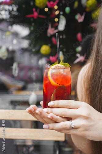 Cold berry lemonade with ice, mind and lemon in female hand against the summer outdoor cafe