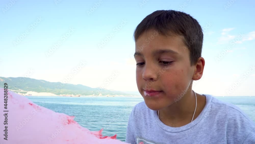 The boy is eating cotton candy on the seashore. Pink cotton candy. Dark hair on the head. Dark skin. Blue sky. Dark water. Close-up. The boy is eating cotton candy on the seashore. Pink cotton candy. Dark hair on the head. Dark skin. Blue sky. Dark water. Close-up.