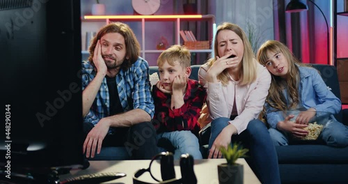 Likable relaxed calm family sitting on the couch at home in the evening and watching bored movie or uninteresting show on TV,mother yawning,daughter eating pocorn,dad and son putting their heads on
