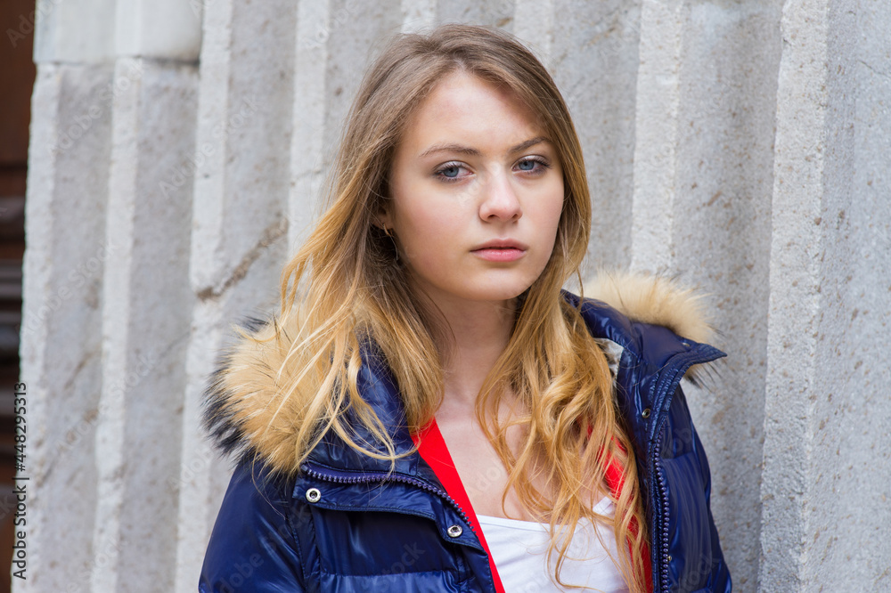 Dressing in a blue Down hooded jacket, a blonde teenage girl is standing outside, thoughtfully looking at you.. Dressing in a blue Down hooded jacket, a blonde teenage girl is standing outside, thoughtfully looking at you..