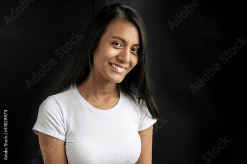 vertical medium close-up of a smiling beautiful black haired latin woman wearing a white t-shirt on a black background with copy space.