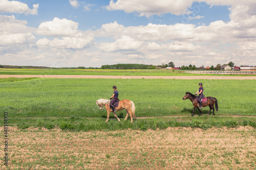 Two horse riders on a Palomino horse and a Dark Bay Horse moving across the beautiful farm field during the daytime. Aerial view of the beautiful meadows. Picturesque cloudy sky in the background.