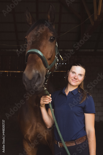 Horsewoman posing with her seal brown horse in the stable. Girl holding the stallion's rope and smiling at the camera. Bonding between human beings and horses concept. © CameraCraft