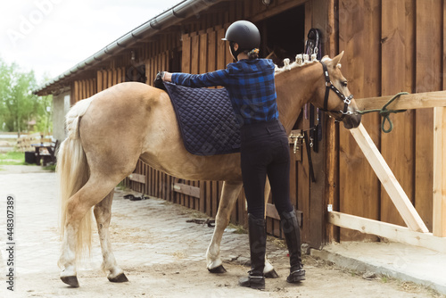 Female jockey saddling up her light brown horse. Placing a saddle pad on the back of her horse to protect it from saddle sores. The girl wearing a helmet and gloves. Preparing for the competition.