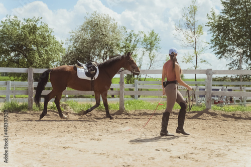 Horsewoman training her dark brown horse during the daytime. Horse routine lunging exercises. Running along the wooden fence in the sandy arena. Low angle shot. Cloudy sky in the background. © CameraCraft