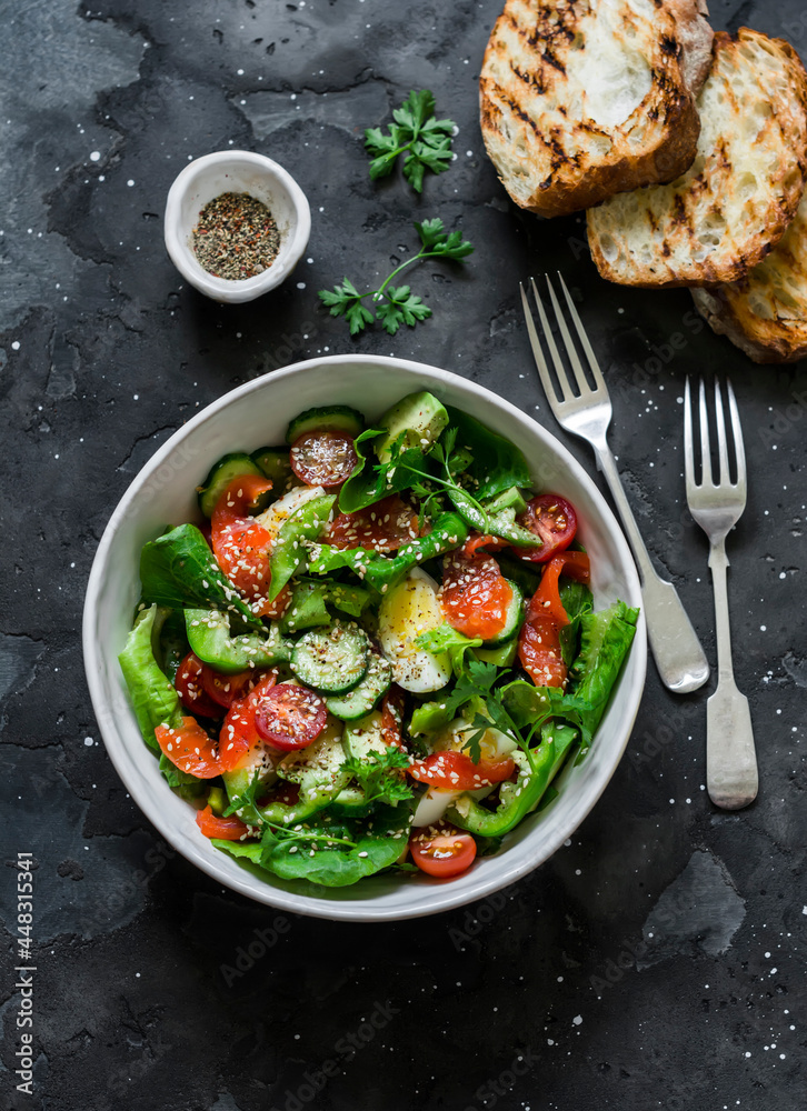 Delicious Mediterranean style salad with salmon, egg, vegetables, lettuce and grilled bread on a dark background, top view
