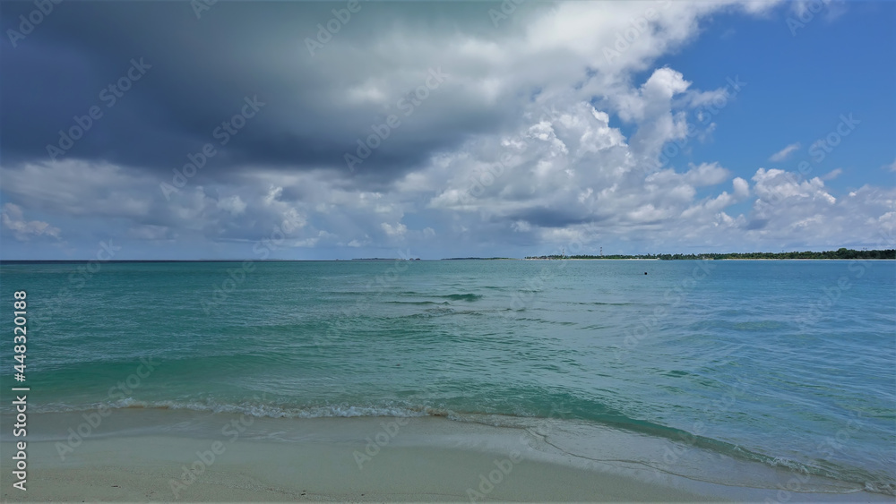A dark cloud hung over the aquamarine ocean among the white clouds. Surf waves splash on the sandy beach. Maldives waiting for rain