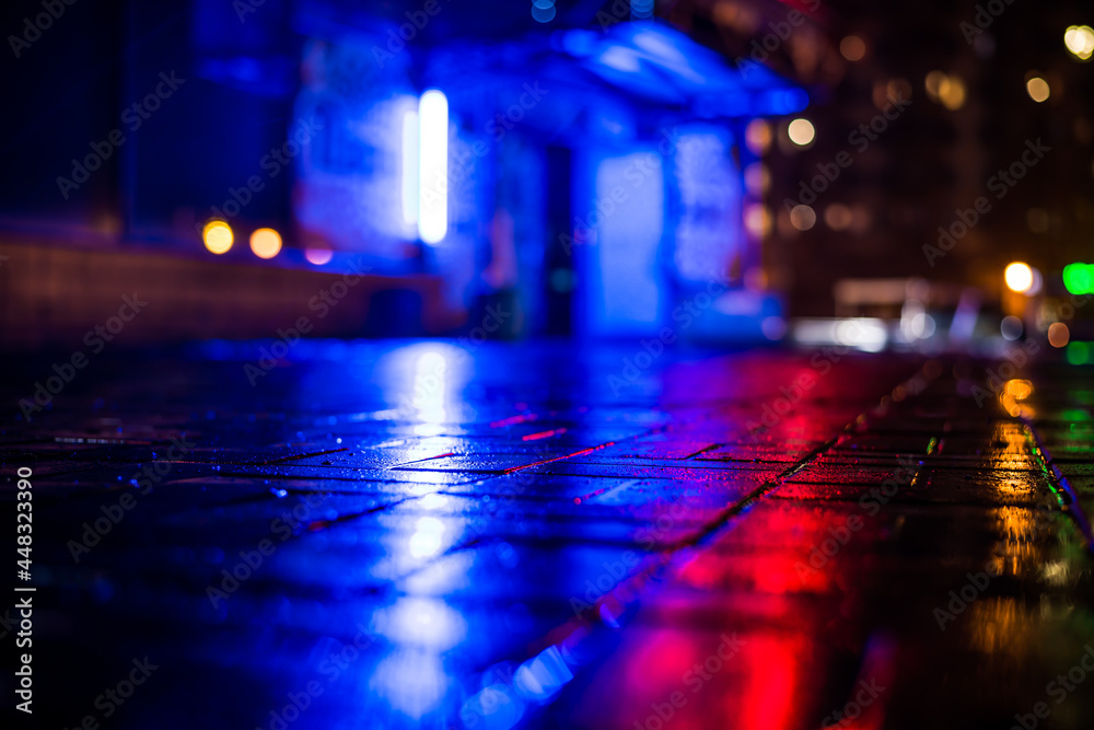 Rainy night in the big city, light from the night club and the windows of the house is reflected in the asphalt. View from the sidewalk level paved with bricks