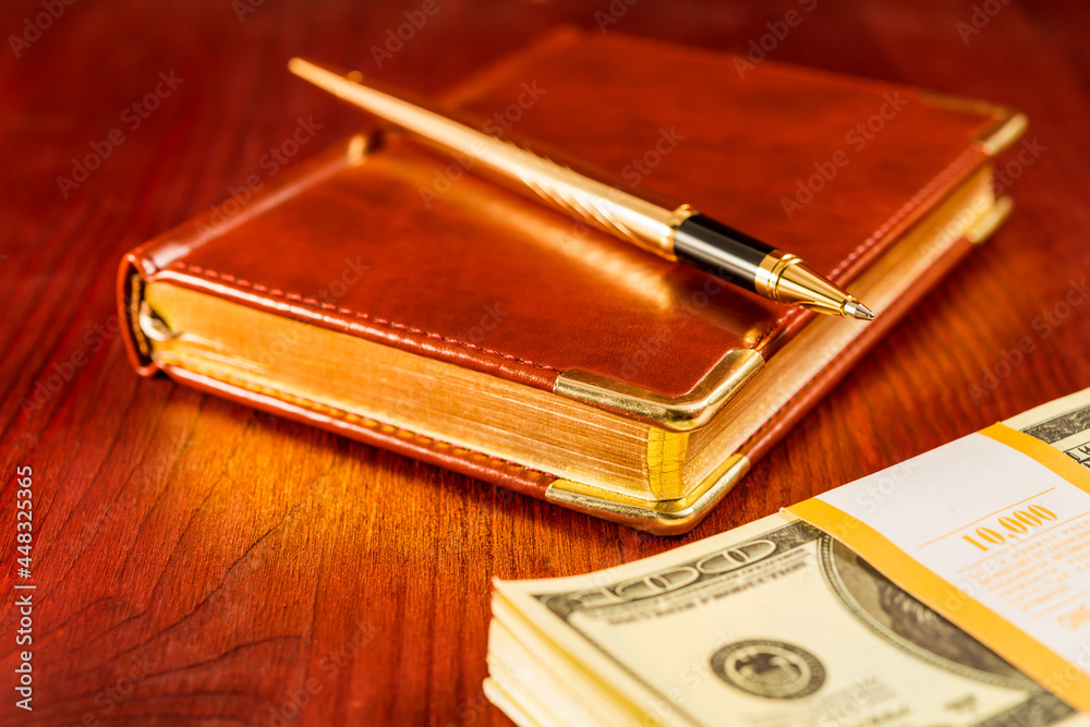 Golden pen on a leather diary with pack of dollars on a mahogany table Golden pen on a leather diary with pack of dollars on a mahogany table