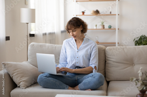 Beautiful young woman sitting crossed legs on comfort sofa with computer on laps, typing message in social network, web surfing information online, choosing goods shopping in internet store.