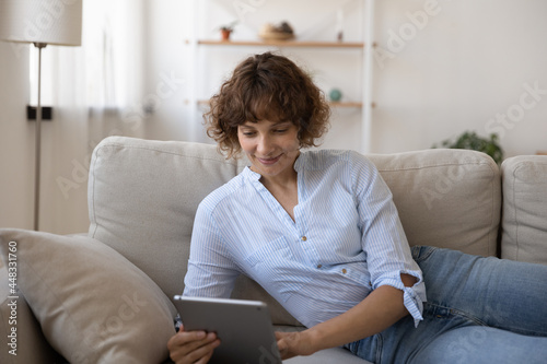 Happy young 35s woman using digital computer tablet, relaxing on comfortable sofa, enjoying playing online game, web surfing information, communicating in social network or shopping in internet store.