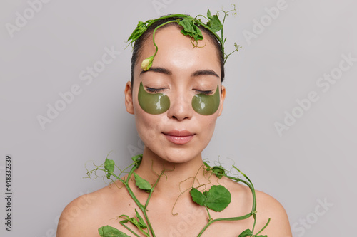 Fototapeta Naklejka Na Ścianę i Meble -  Asian beauty woman cares about skin undergoes facial and body treatments keeps eyes closed applies patches under eyes poses with naked body with green plant around isolated on grey studio wall