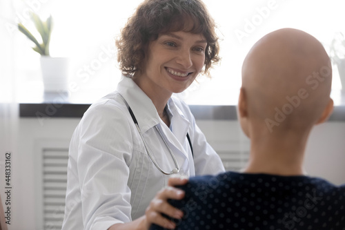 Happy young compassionate kind female oncologist doctor supporting bald after chemotherapy patient woman, telling effective treatment good news or giving psychological support at checkup meeting. photo