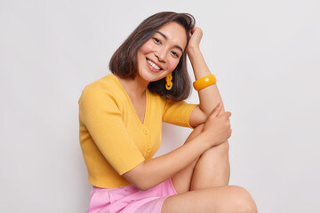 Beautiful teenage girl with eastern appearance leans on hand smiles pleasantly wears yellow jumper pink skirt gazes at camera with cute face expression looks sensual isolated over white background
