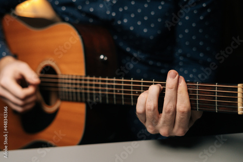 Closeup front view of unrecognizable guitarist male playing acoustic guitar sitting at desk in dark living room, selective focus. Creative musician enjoying leisure activity in apartment.