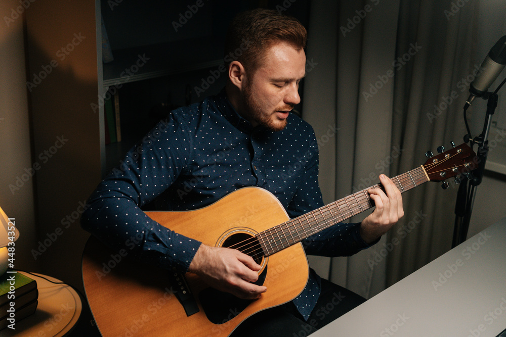 Obraz premium Talaented guitarist singer man emotionally singing into condenser microphone and playing acoustic guitar in dark home recording studio. Creative musician leisure leisure in apartment