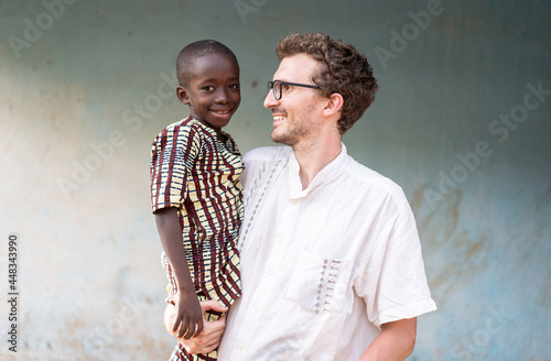 Profile view of a young caucasian man carrying a smiling little African orphan in traditional outfit looking in camera; international adoption concept photo
