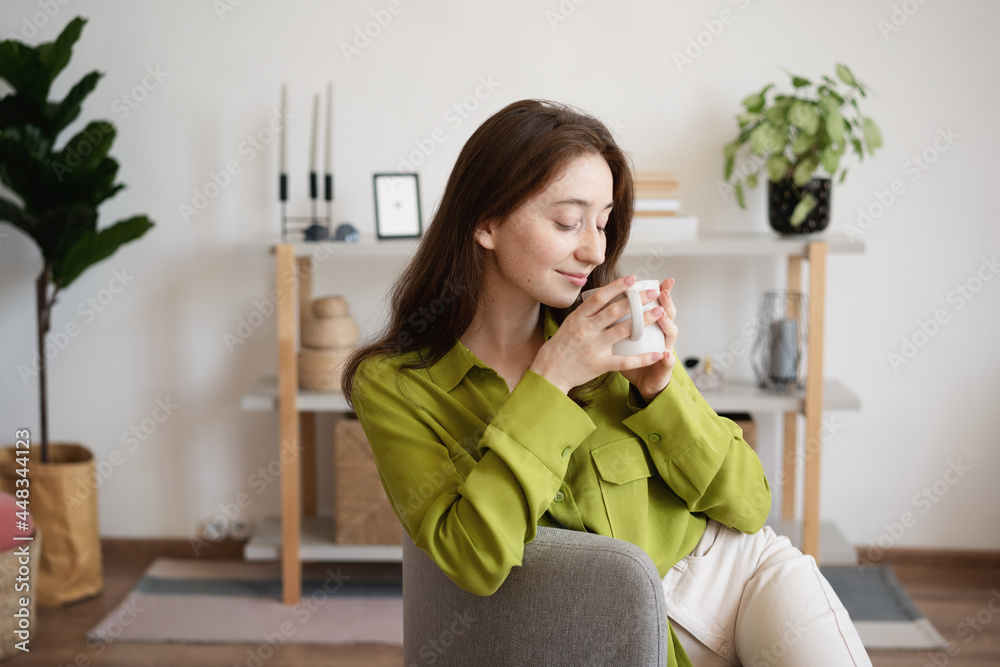Young beautiful smiling woman in a green blouse with a mug of coffee sitting at home on a gray armchair in the living room Young beautiful smiling woman in a green blouse with a mug of coffee sitting at home on a gray armchair in the living room