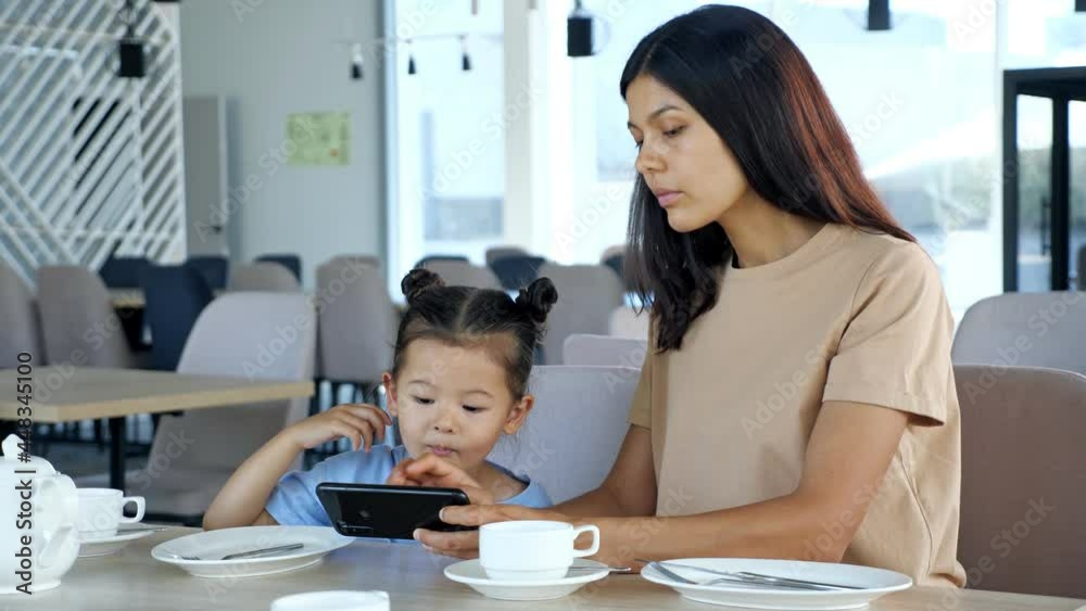 Lady shows daughter smartphone. Asian woman brunette and small kid girl watch video on black gadget sitting at restaurant table waiting for order close view Lady shows daughter smartphone. Asian woman brunette and small kid girl watch video on black gadget sitting at restaurant table waiting for order close view