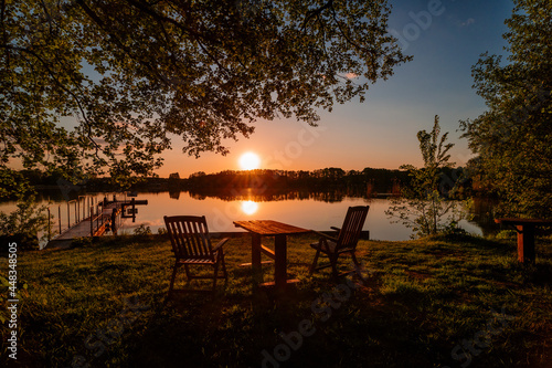 Ein Sonnenuntergang am See und im vordergrund steht ein Tisch und zwei Stühle. Romantische Stimmung