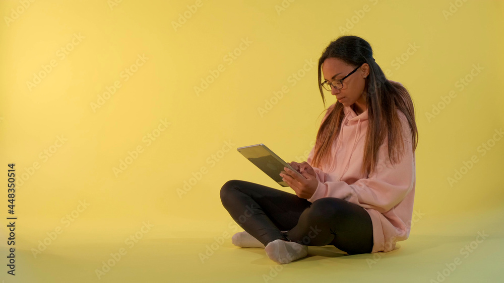 Attractive mixed-race girl reading something on tablet and then looking over her glasses to the camera. Yellow background.