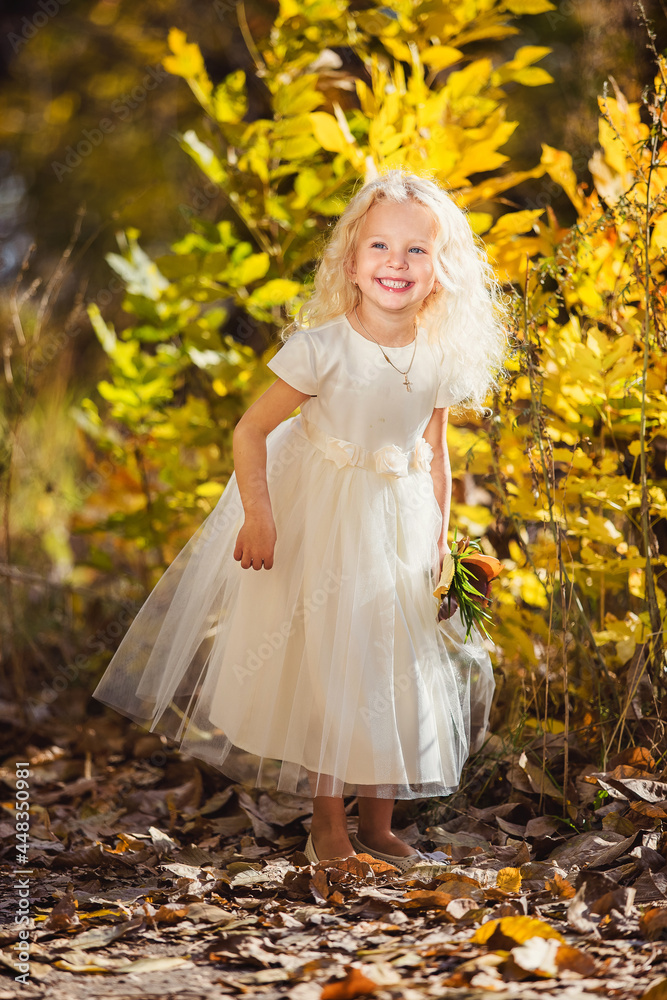 A little girl in white dress who smiles on a background of yellow leaves