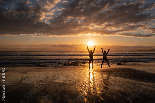 Two people in silhouette jumping in the air at Milford beach at sunrise, Auckland