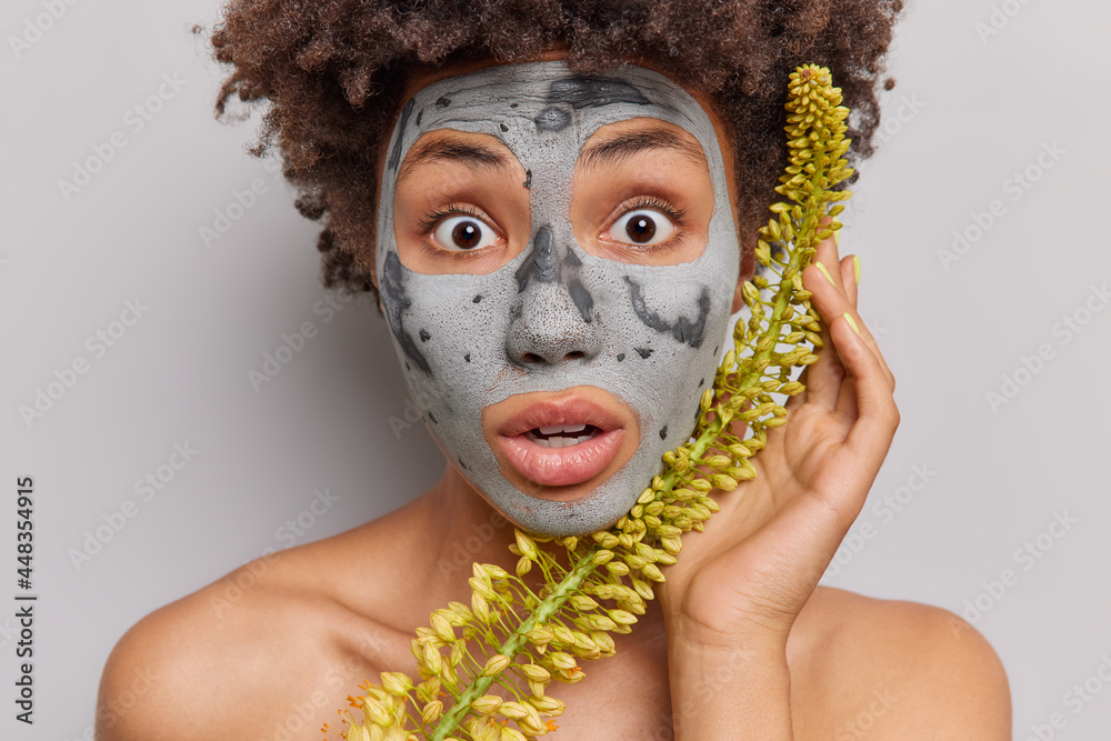 Close up portrait of surprised ethnic curly woman stares impressed at camera applies clay herbal mask cannot believe her eyes stands topless indoor against grey background. Natural skin care