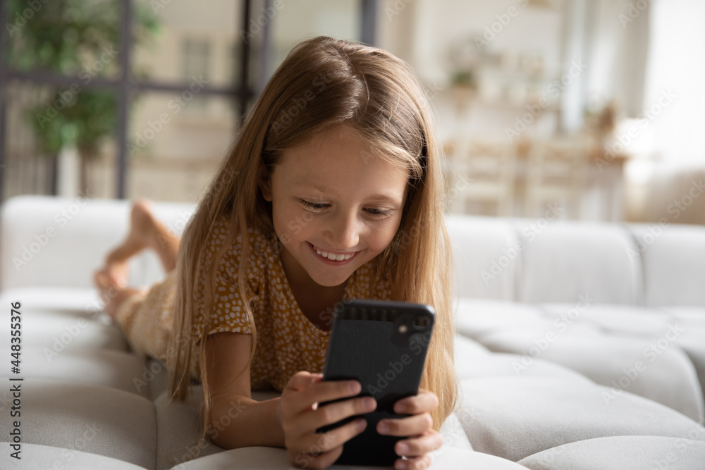 Close up adorable smiling little girl using smartphone lying on couch alone, happy preschool child kid spending leisure time with modern device, looking at screen, watching cartoons, playing game Close up adorable smiling little girl using smartphone lying on couch alone, happy preschool child kid spending leisure time with modern device, looking at screen, watching cartoons, playing game