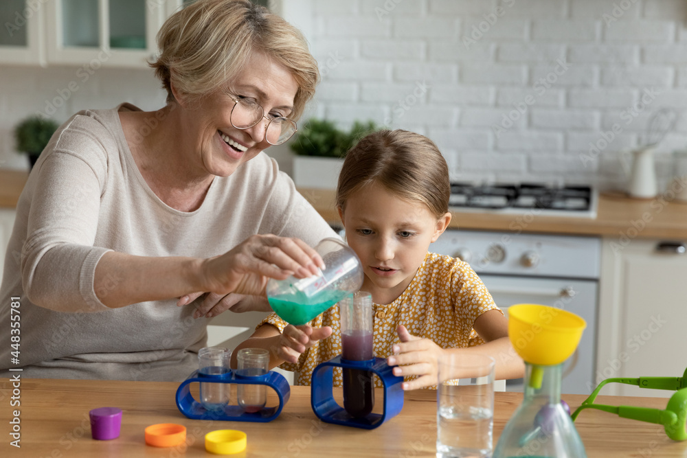 Happy mature woman in glasses with curious little girl making funny experiments in kitchen, smiling aged grandmother and adorable granddaughter having fun with toy laboratory, educational activity Happy mature woman in glasses with curious little girl making funny experiments in kitchen, smiling aged grandmother and adorable granddaughter having fun with toy laboratory, educational activity