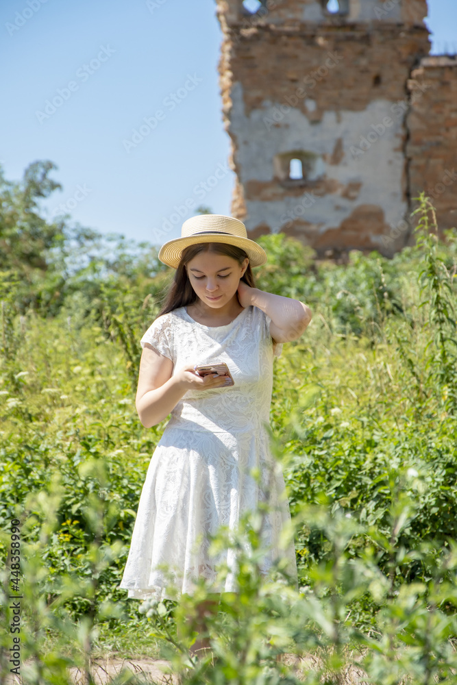 A young girl of 17-20 years old in a straw hat and a white dress looks at a mobile phone against the background of an old ruined castle overgrown with grass.
