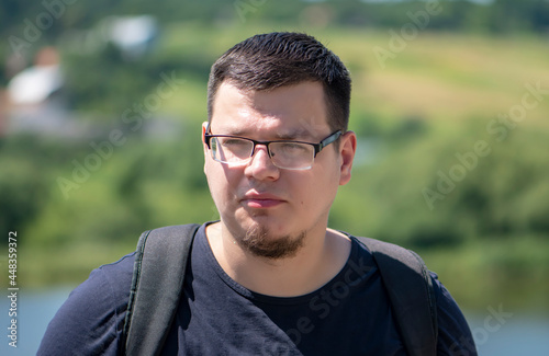 Street portrait of a serious young man 25-30 years old in glasses with a backpack against the background of nature and the lake. Perhaps he is a tourist or a traveler, an IT specialist on vacation.
