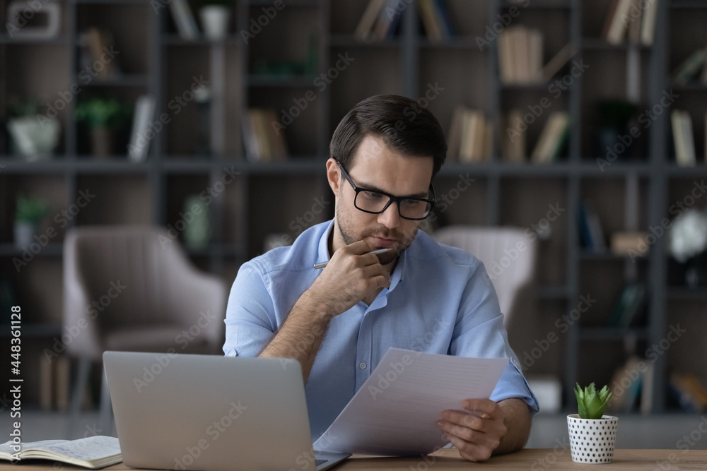 Working with papers. Serious millennial male entrepreneur work at home office alone hold read document hardcopy think on conditions terms. Focused young man engaged in paperwork edit text of agreement - obrazy, fototapety, plakaty 