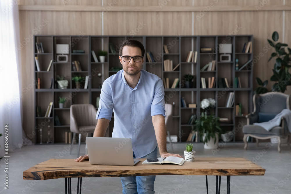 Portrait of young handsome man teacher mentor trainer in glasses stand in classroom lean on work desk with laptop phone paper daybook. Casual guy employee look at camera pose by home office workplace Portrait of young handsome man teacher mentor trainer in glasses stand in classroom lean on work desk with laptop phone paper daybook. Casual guy employee look at camera pose by home office workplace