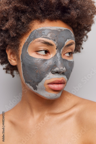Close up portrait of young Afro American woman with curly hair looks away pensively applies nourishing clay mask for skin rejuvenation reducing black heads and pores stands bare shoulders indoor