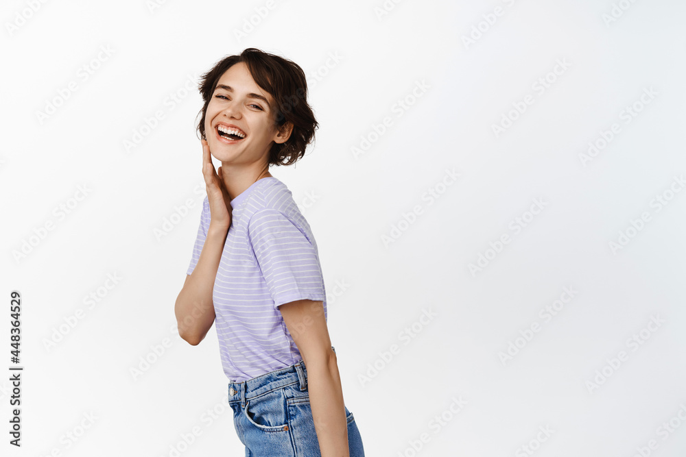 Image of candid beautiful girl with short pixie hairstyle, no makeup, touching her clean healthy face, laughing and smiling happy, white teeth, posing in casual clothes against studio background Image of candid beautiful girl with short pixie hairstyle, no makeup, touching her clean healthy face, laughing and smiling happy, white teeth, posing in casual clothes against studio background