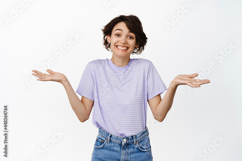 Nothing to say sorry. Smiling cute and coy brunette woman spread empty hands sideways, shrugging shoulders silly, acting as if dont know anything, stanidng over white background photo