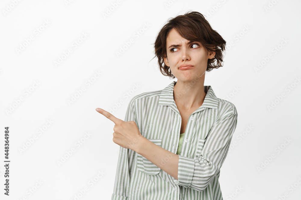 Skeptical, suspicious young woman pointing and looking left with raised eyebrow, being unsure, having doubts, standing against white background Skeptical, suspicious young woman pointing and looking left with raised eyebrow, being unsure, having doubts, standing against white background