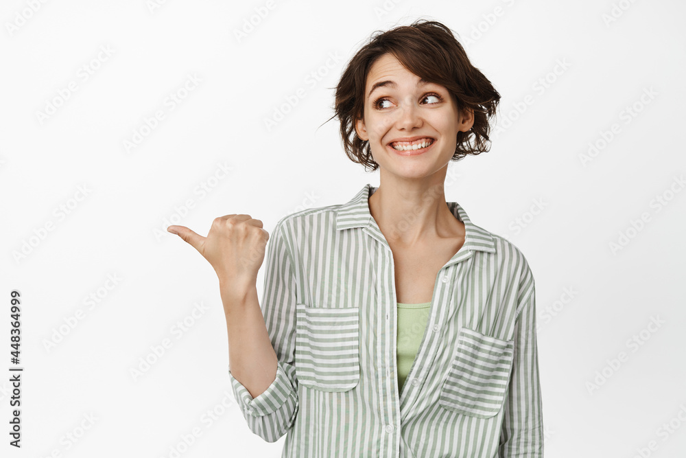 Portrait of cheerful smiling woman pointing, looking left at copy space, showing advertisement, wearing casual shirt, white background Portrait of cheerful smiling woman pointing, looking left at copy space, showing advertisement, wearing casual shirt, white background