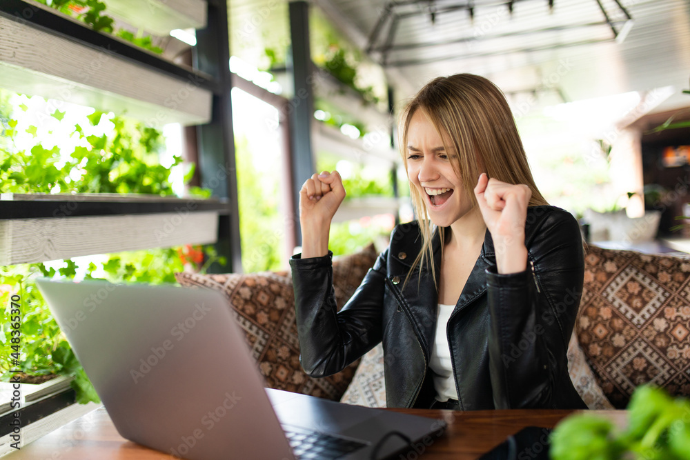 Girl rejoices in winning having raised her hands up sitting in front of laptop.Girl sitting in cafe, watches online sports match on computer. Girl rejoices in winning having raised her hands up sitting in front of laptop.Girl sitting in cafe, watches online sports match on computer.