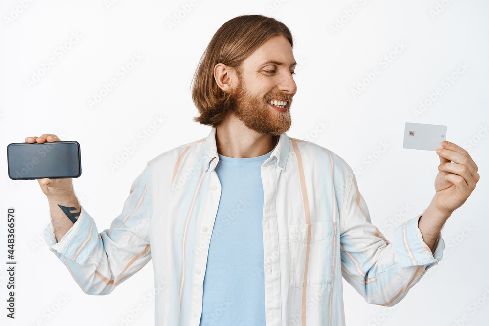 Image of handsome guy holding credit card, smartphone horizontally, smiling and looking at card with happy face expression, white background Image of handsome guy holding credit card, smartphone horizontally, smiling and looking at card with happy face expression, white background